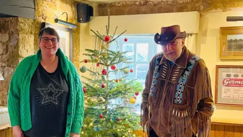 Garnett family A woman in a green jumper and a man in a cowboy outfit smile in front of a Christmas tree.