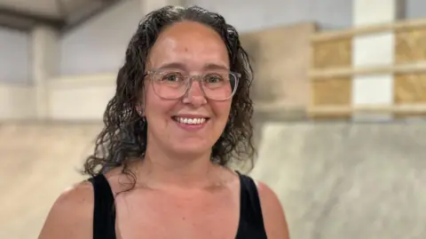 Aimee Dexter/BBC Rochelle is standing in front of skate ramps. She is off centre to the left and has long curly brown hair. She is wearing clear framed glasses and a black vest top, and is smiling.