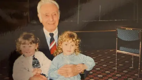 Mark Barnes Sir Stan Matthews with white hair, blue blazer, light and white striped shirt with a white collar and a Stoke City tie of red, black and white. He is with two small children.
