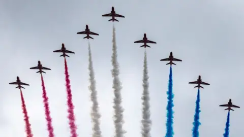 Nine Red Arrows aircraft fly against a grey sky, trailing smoke coloured red, white and blue behind them.