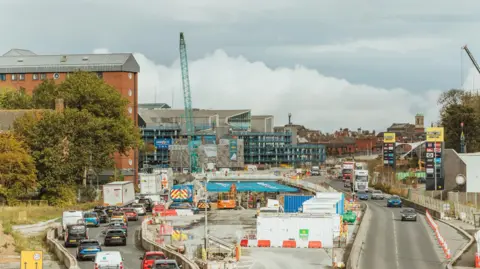 Traffic making its way through a contraflow system in Hull city centre, with machinery, a crane and traffic cones in view. 