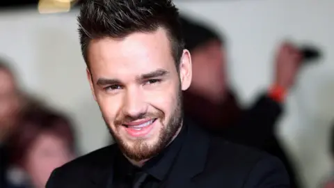 Singer Liam Payne, dressed in a black suit, shirt and tie, poses for photographers at a movie premiere.