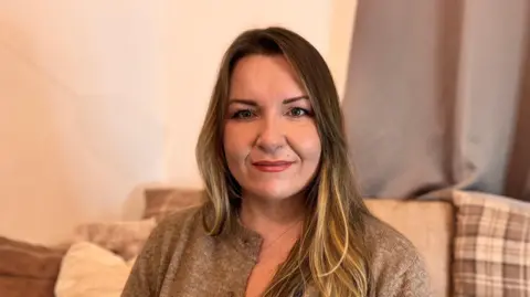 Sonja Leason in her living room wearing a biscuit coloured cardigan, long brown hair with highlights. 