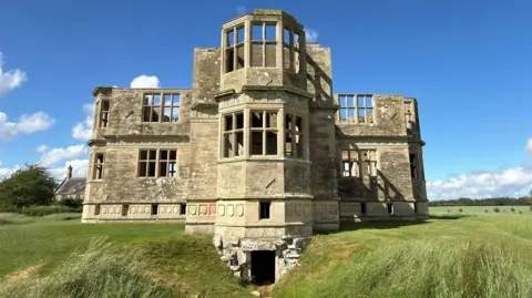 Lyveden Lodge, a roofless late 16th Century building built out of cream-coloured stone. It was built in the shape of a cross and the view shows three of its four sides, with two sides on either side of a third which juts out in the middle. It is surrounded by grass and above is blue 