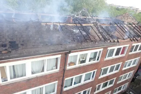 London Fire Brigade Damaged roof of black of flats with smoke rising
