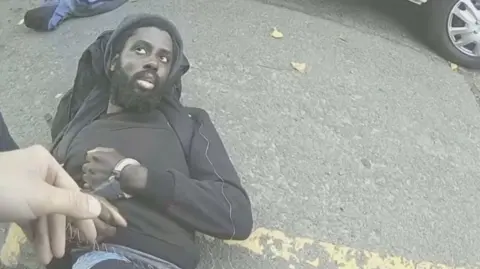 Nottinghamshire Police A still taken from police body cam footage of Valdo Calocane being arrested. He is on the floor wearing all black clothing and in handcuffs. He is looking up at a police officer and appears expressionless. 