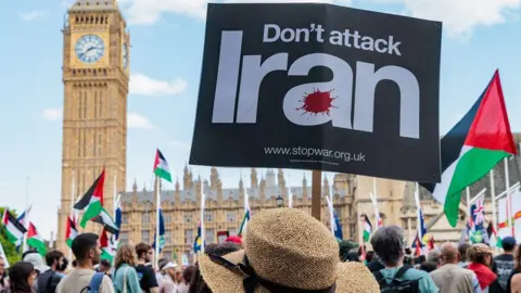 A crowd of people face the Houses of Parliament. In the foreground is a person wearing a straw hat and holding a sign that reads: Don't attack Iran