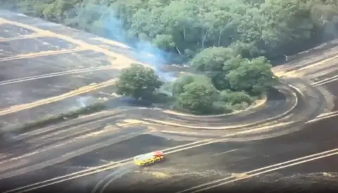Drone footage of a blackened field following a fire, with burned crops and some smouldering smoke. There is a fire engine in the field and some trees in the middle.