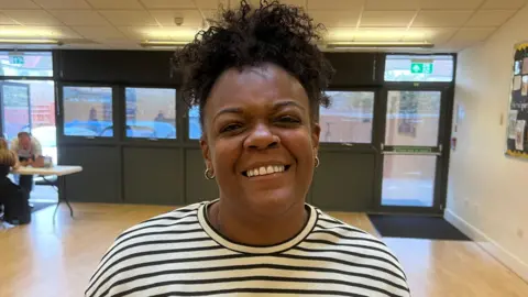 Simon Dedman/BBC Sharleen Dacosta, a woman who is standing in a hall with wooden floors. She is looking directly at the camera and smiling and is wearing a black and white striped T-shirt. 