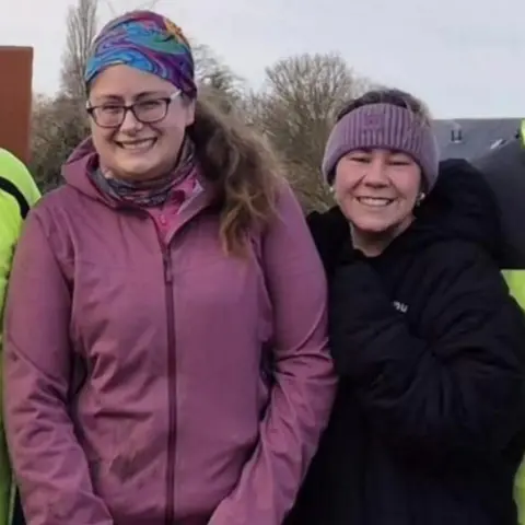 Supplied Two women are standing and smiling and both wearing coats. On the left the woman is wearing a blue bandana and glasses and has long brown hair tied back, she is also wearing a pink coat and blue trousers. On the right is a woman in a black coat and a purple headband.