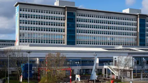 University Hospital Wales main building seen from the front outside. A large high-rise rectangular building rises up behind and a two-storey building is visible in front 