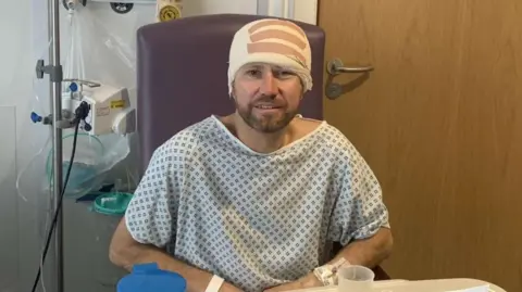 Brain Tumour Research Nick Ball wearing a blue and white patterned hospital gown and a large bandaged around his head. He is sitting in a purple chair in a hospital room, bedside medical equipment and a wooden door. He is looking at the camera but looks weak and unstable.