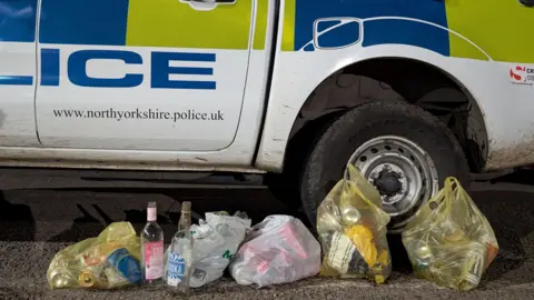 North Yorkshire Police Five carrier bags of rubbish are on the ground by a police car, three yellow bags and two white ones. They are full of cans and glass bottles, with two glass bottles sitting in front of one of the bags. 