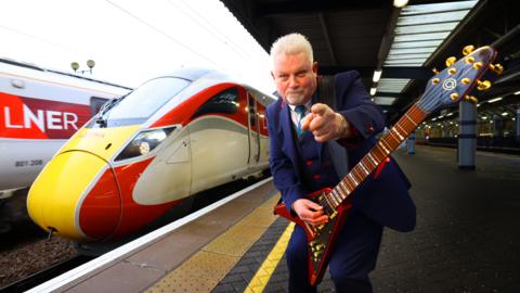 Robb Weir holding a guitar with LNER train in the background at Newcastle Central Station 