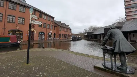 Coventry's Canal Basin has water surrounded by red brick buildings. A signpost shows directions to Brunston. In the foreground a statue of James Brindley. Canals can also be seen on the water. 