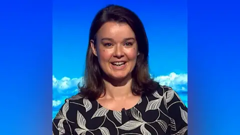 Gillian Smart has shoulder-length brown hair, wears a black top with white leaves pattern
