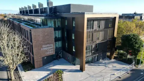 An aerial shot of the exterior of Worthing Integrated Care Centre building shows a large modern building with a sign for the centre and a paved area i front, surrounded by trees.