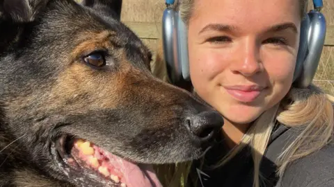 Thin Blue Paw Foundation A selfie-style image of Carleigh-Jay Callaghan and Bane the dog. Carleigh, on the right of the image, is wearing silver headphones and a grey top. She is looking into the camera. Bane, a German Shepherd-type dog, has his mouth open and is looking away from the camera.