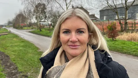 Claire Dean has shoulder length blonde hair parted in the middle, she is wearing a woollen beige and black scarf over a black puffer coat. Behind her is a single track road with some buildings visible and grass verges on either sidet with some trees growing.