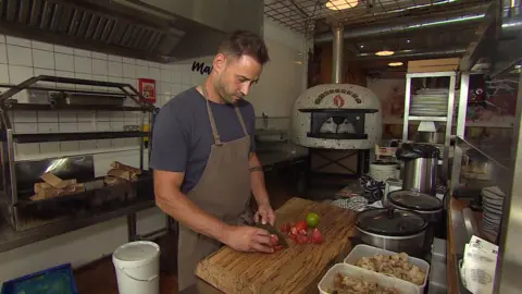 A man with brown hair is in a pizza kitchen. He is cutting red tomatoes on a wooden chopping board and there is a wood fired pizza oven in the background. The man has brown hair and is wearing a navy t-shirt under a brown apron