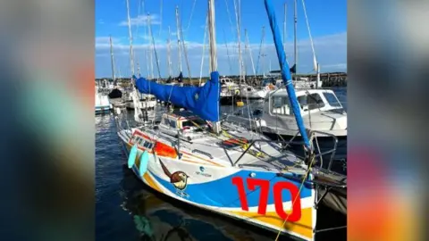 Ella Dawson Foundation Little Wren in the water at a marina. The small sailing boat is white with a blue and yellow stripe and the number 170 in red on the side. It also has a drawing of a bird on the side of it.