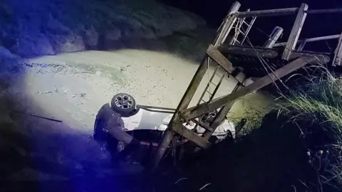 HM Coastguard A car is on its side in a creek, surrounded by mud, at nighttime