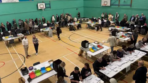 people are standing around a sports hall with people sitting at table and chairs counting votes.
