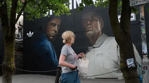 PA A mural of Oasis on a wall - Noel is wearing a denim shirt and Liam is wearing a buttoned light grey shirt. A woman, wearing a short sleeved grey t-shirt looks up at the mural as she passes.
