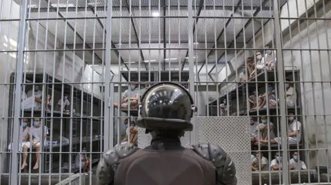 A guard stands with his back to the camera in front of a jail cell in Cecot, in which dozens of prisoners are visible 