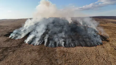 An aerial picture of a large mountain fire actively burning in a large circular shape. Lots of grey smoke is rising into the sky, leaving a blackened patch on the ground. 