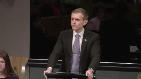 Senedd Cymru Joe Martin speaking at a lecturn in the Senedd, while a colleague sat to the left of him looks down.