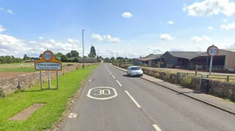 Google The B6412 road leading into Temple Sowerby. A sign on the right states the name of village as well as a 30 mph speed limit. A farm building stands on the right.