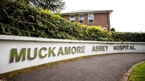 PA Media A white wall with silver letters on it reading "Muckamore Abbey Hospital". There is a black tarmac path in front of it. Behind it is a hedge and the second storey of a two-storey red brick building can be seen.