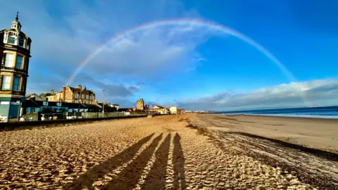 Kath Thomson A rainbow over a sandy beach. There are houses and buildings on the left and the sea on the right. The shadows of four people are on the sand. There are a few clouds in the bright blue sky.