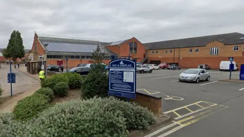 A council car park in Market Harborough next to the town's market hall. A number of cars are parked in spaces, and there is a shrubbery bed just outside the car park, and a large Pay & Display sign can be seen.