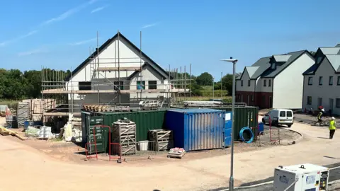 A white two-storey house surrounded by scaffolding is seen on a building site. Blue and green containers can be seen in front of the house, while workers in hi-vis jackets in the distance are walking near houses to the right that look complete