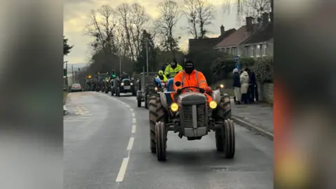 Somerset Tractor Runners A line of tractors drive down a residential street. A group of people watch them pass. The tractor drivers are wearing high vis jackets.