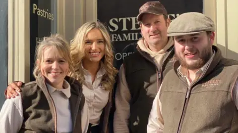 Two blonde women and two men with brown hair and flat caps stand outside a bakery branded "Steele's Family Bakers". All four are wearing brown fleece gilets with the bakery branding