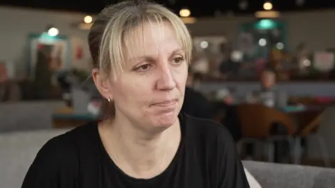 A woman looks slightly off camera. She is sitting on a sofa, inside a café. She has blonde hair and brown eyes and wears a black shirt.