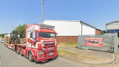 A red Scorpion Engineering truck outside a gate with a sign which says 'Scorpion Trailer Service Centre' - there are two buildings with corrugated wall panels.
