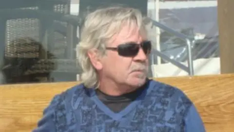 Michael Shirley has collar-length grey hair and is wearing black sunglasses and a blue V-neck jumper over a black T-shirt. He is looking to the side while sitting on a wooden bench.