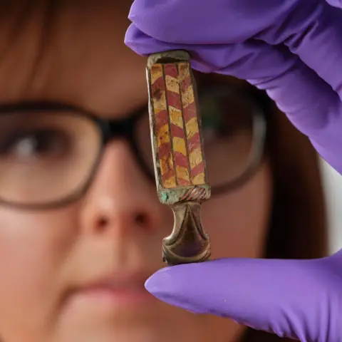 PA Media A woman wearing glasses holds up the brooch in a hand while wearing purple, protective gloves. The brooch is a a rectangle shaped object with a brown taper shaped end. The rectangle shaped section is decorated with a red and yellow chevron-like pattern.
