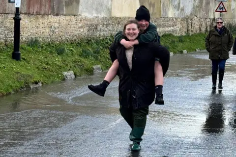 Hannah Crawford The rector Leila Mathers is wearing greed waders as she carries a schoolchild in a piggy back across a flooded road.