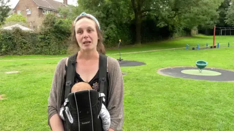 Lauren Slater in a brown cardigan with a baby sling attached stands in front of a park