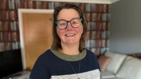 Ceri Walsh is standing in her living room. She is wearing a jumper that is navy, grey and dark green. She also has a necklace. Ceri is also wearing blue-framed glasses and is smiling at the camera.