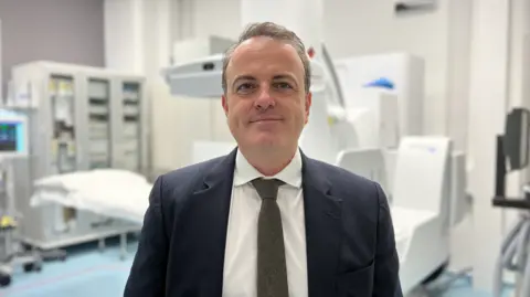 Dr Nolan Walker stands in a clinical room. Behind him is medical equipment, storage cabinets and a hospital-style bed. He is wearing a dark business suit, white dress shirt, and a dark tie.