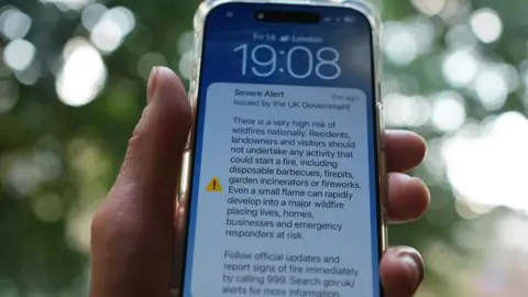 A hand holding an iPhone which includes a notification of an emergency alert from the UK Government on wildfires.