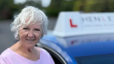 Olwen has white short hair. She smiles for the camera in front of her learner car. She is wearing a pink sweater.