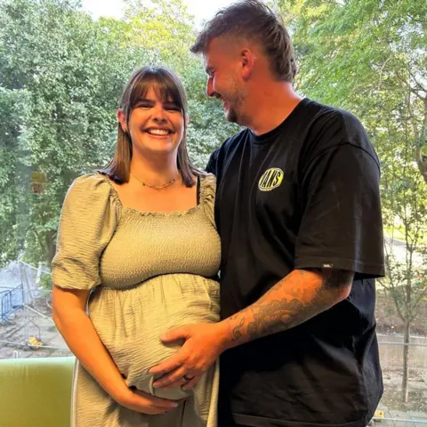 A pregnant woman in a light green dress with shoulder length brown hair and a fringe is smiling at the camera and stood up whilst holding her bump. A man next to her in a black vans top and with arm tattoos and short brown hair has his hand on her stomach and is smiling and looking at her. You can see a window and greenery behind them.