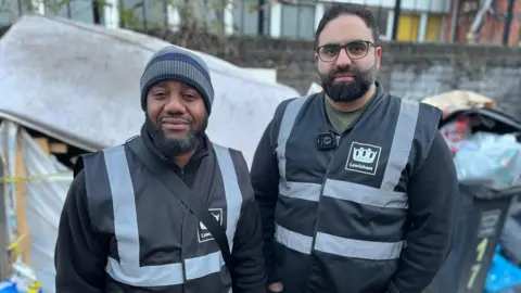 Alain N'Guessan Bi and Eiman Rostami wearing dark Lewisham clothing. They are standing in front of overfilling bins and a stained mattress